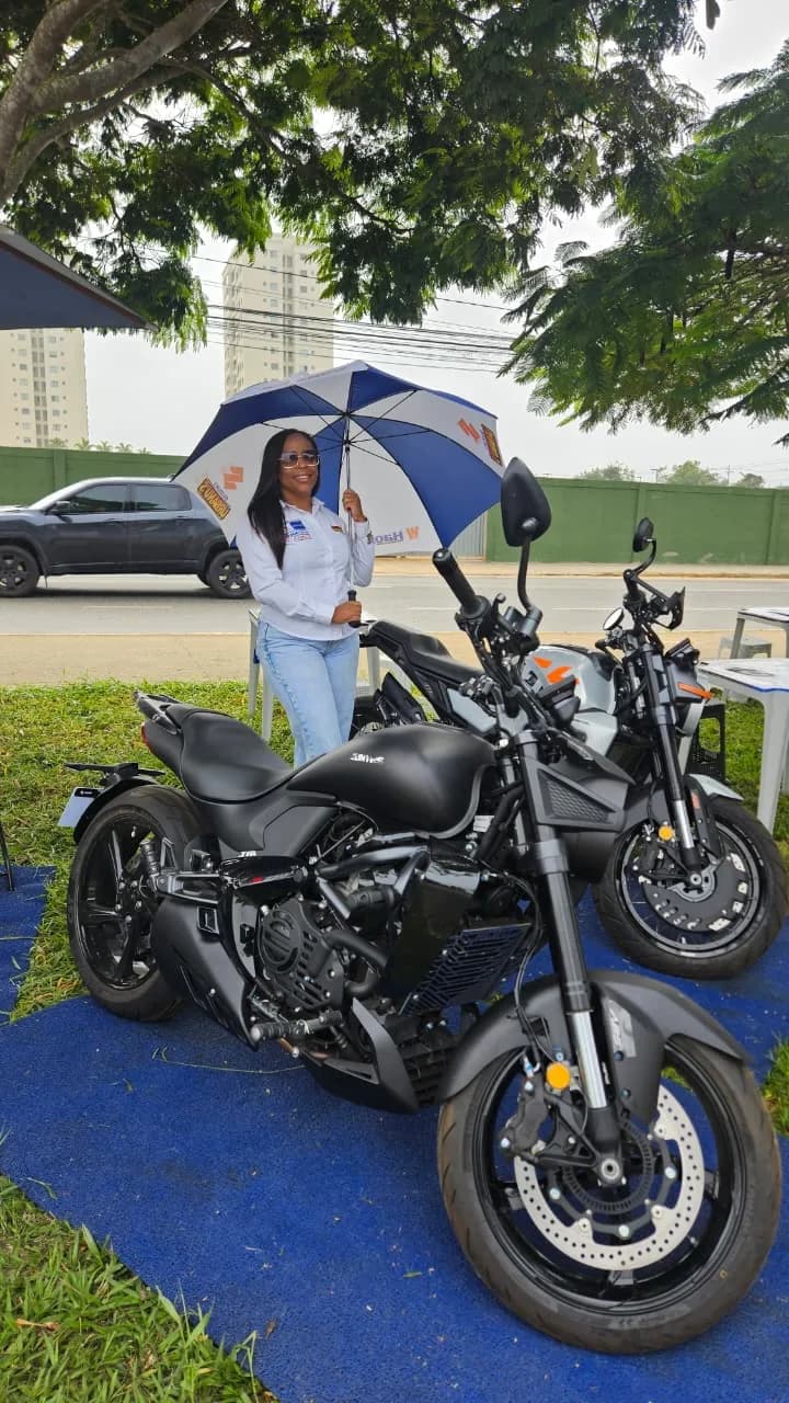 Promotora da Urbanu's com sombrinha ao lado das motos expostas na grama, sobre tapete azul