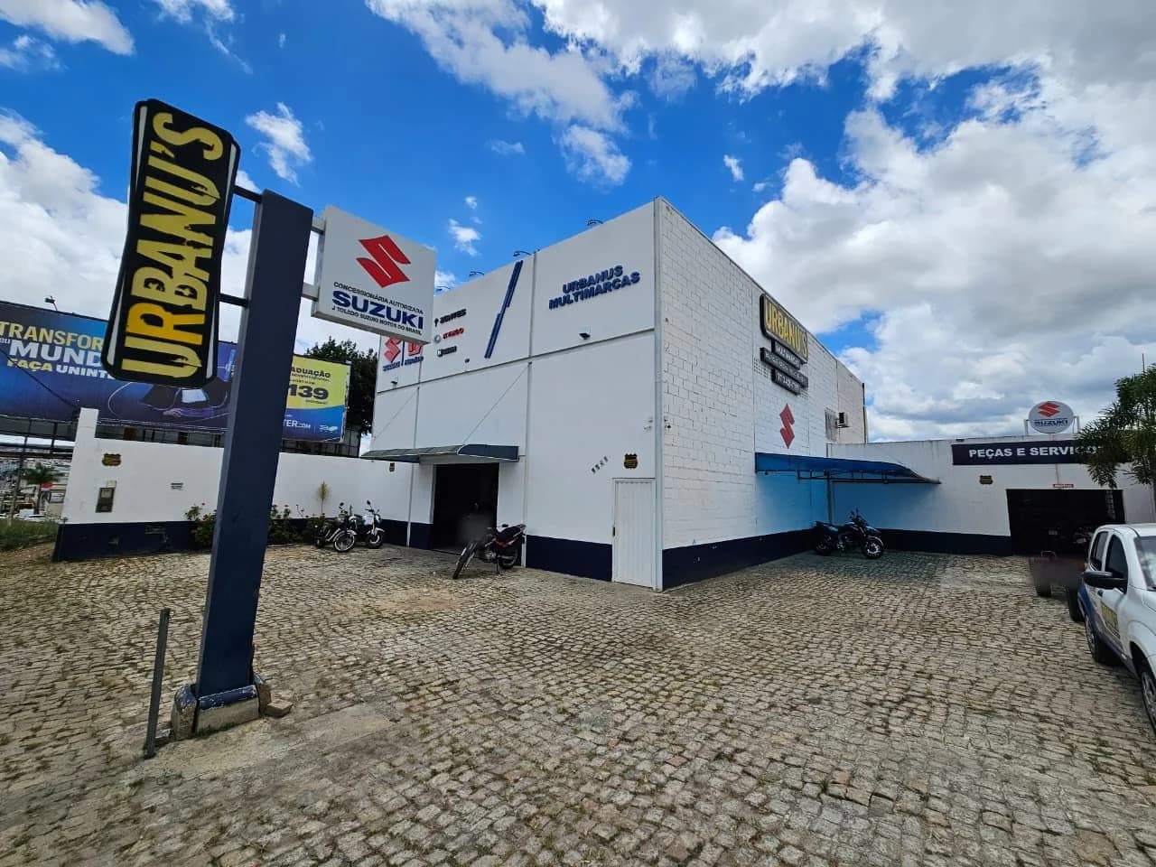 Fachada da Suzuki Urbanu's, com o totem Urbanu's, a placa de concessionária autorizada Suzuki e a entrada de peças e serviços